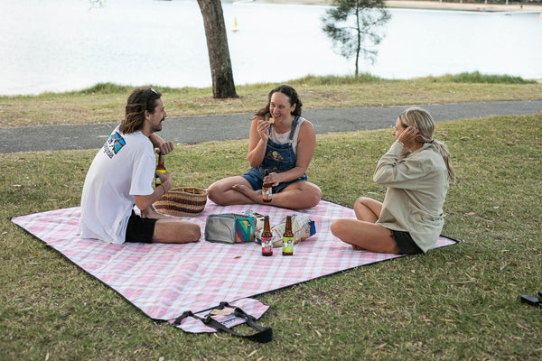 Pink checkered recycled picnic blanket spread on grass with three people sitting and enjoying food and drinks by a lake.