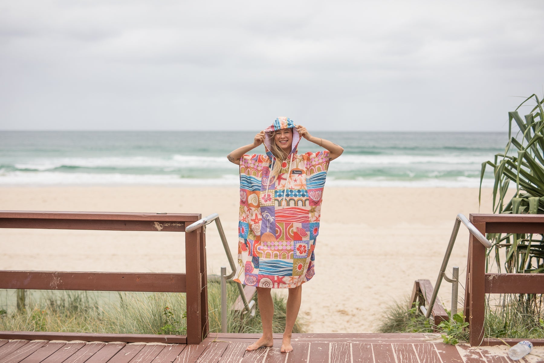 Woman wearing a Nakie Retro Shores hooded towel enjoying ocean views from a coastal lookout.
