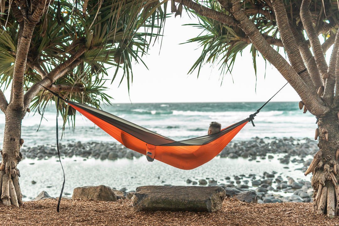 Person relaxing in a Nakie Sunburnt Orange hammock between trees overlooking the ocean.