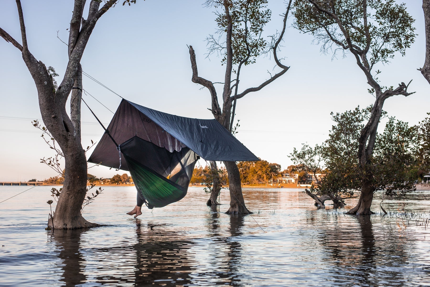 Person relaxing in a Nakie hammock, bug net, and tarp set up between trees over shallow water at sunset.