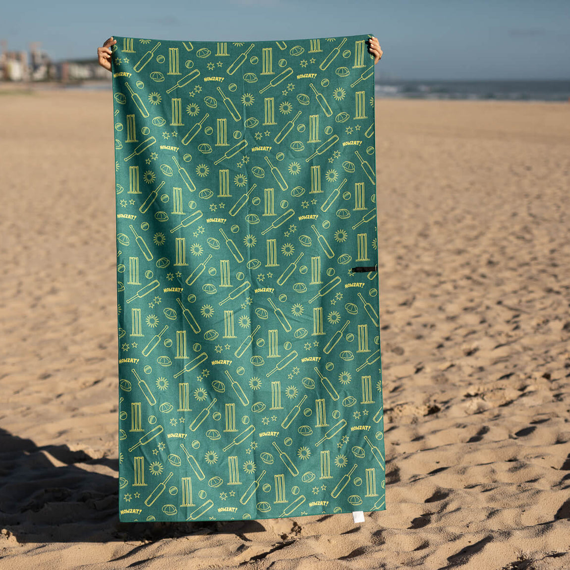 Teal recycled beach towel with cricket-themed gold pattern including bats, balls, and stumps, displayed on sand.