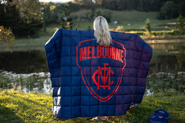 Navy quilted puffy blanket with red Melbourne Demons AFL crest logo, displayed outdoors by water.