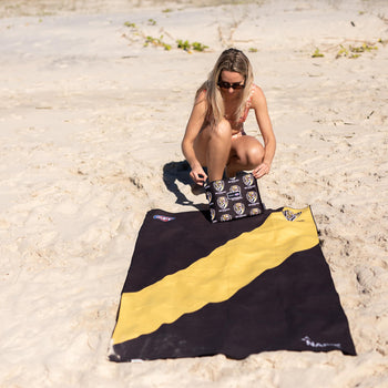 Black and gold Richmond Tigers AFL beach towel with diagonal stripe design, displayed on sand at the beach