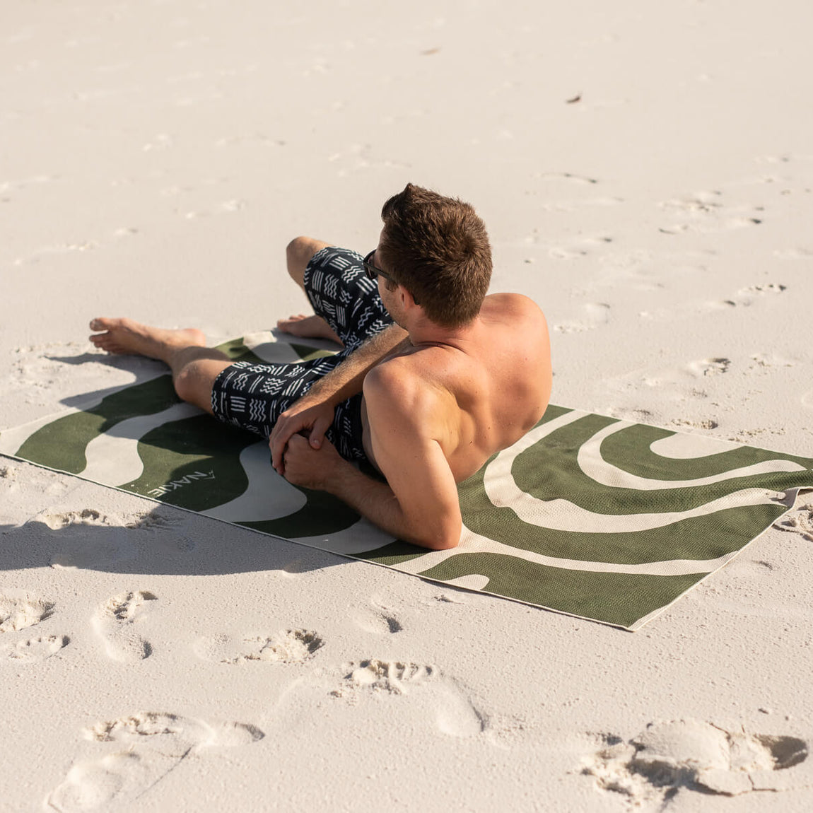 Eucalyptus green beach towel with abstract cream wave pattern, displayed on sand with man and child sitting on it.