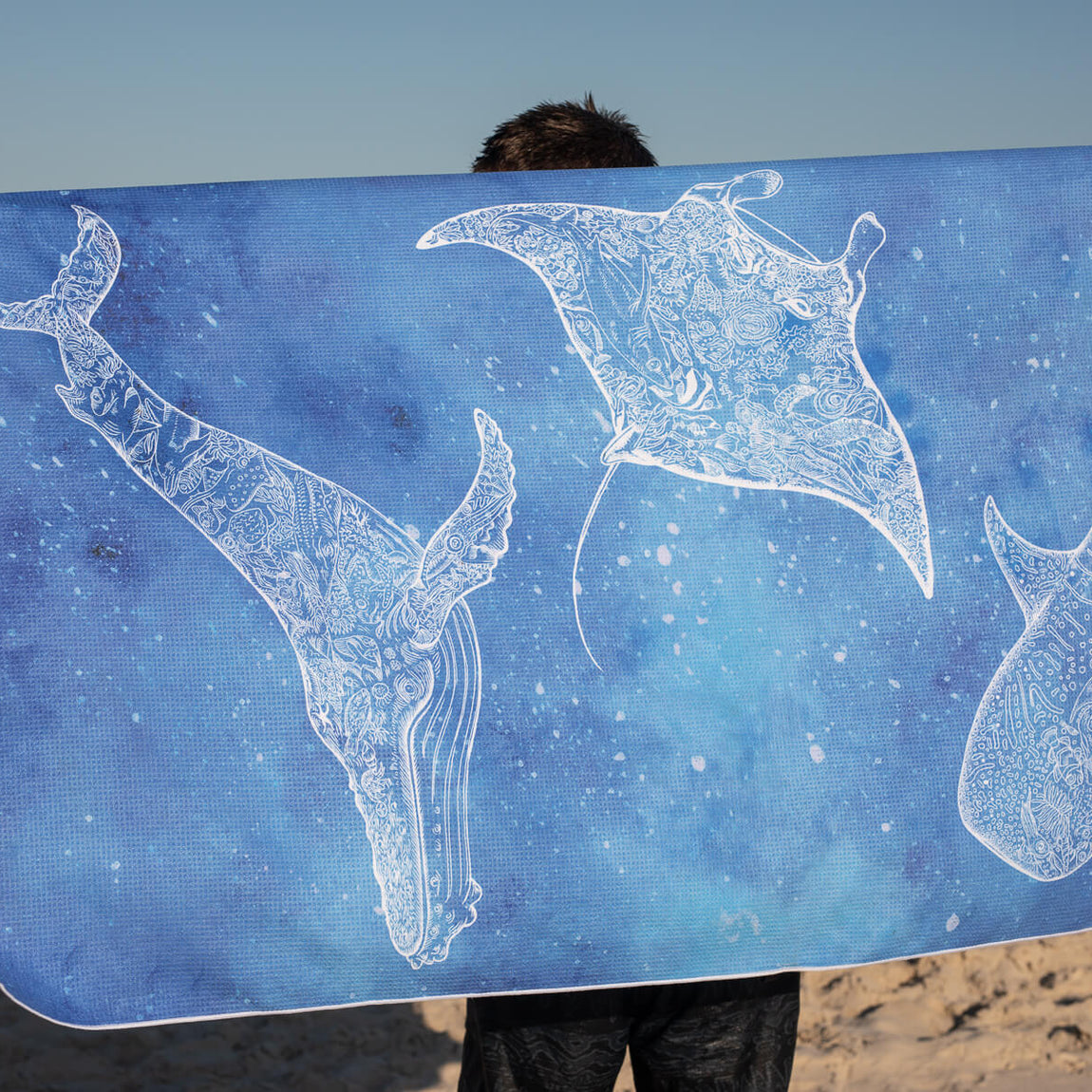 Blue beach towel with white decorative manta ray and stingray illustrations, sand-free texture, held at seaside.