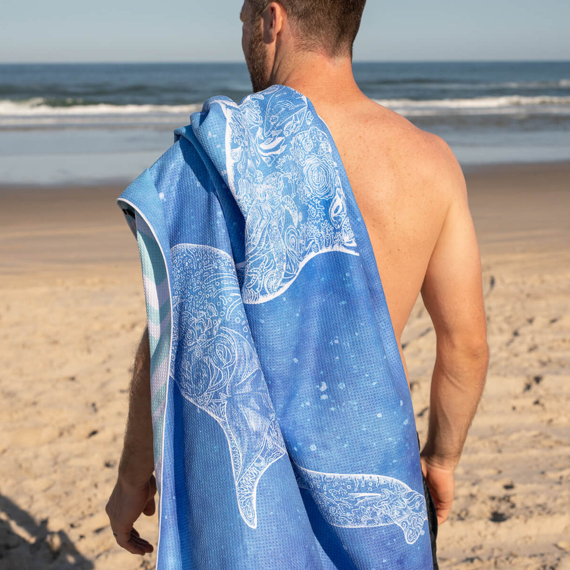 Blue recycled beach towel with white whale and ornamental pattern, shown draped over man's shoulder at beach
