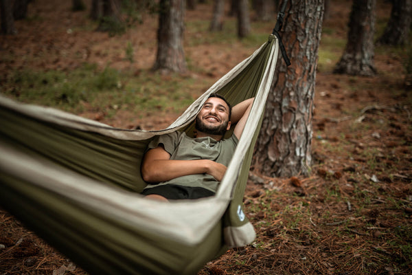 Olive green recycled hammock with black straps suspended between trees, occupied by smiling man in forest setting.