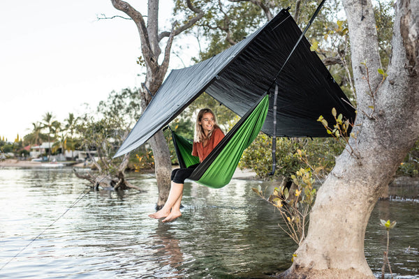 Green and black recycled nylon camping hammock with attached black tarp rain fly, suspended over water between trees.